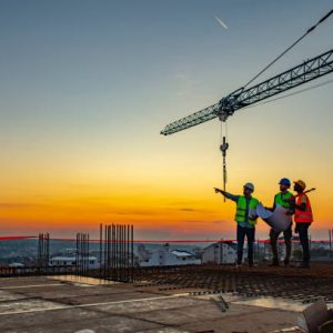 Three Multi-Ethnic construction workers in uniform standing at construction site with crane in background, discussing building plans while holding blueprint at sunset under the tower crane.